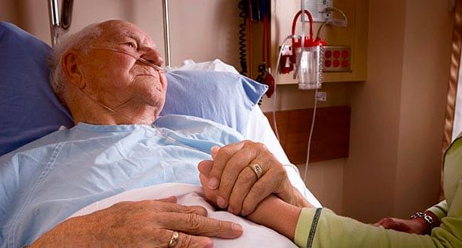 Old man lying in hospital bed holding daugher's hand